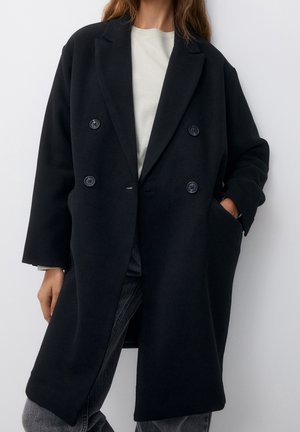 Femme portant un long manteau noir à double boutonnage sur une chemise blanche et un jean gris, une main dans la poche du manteau, debout devant un fond uni.