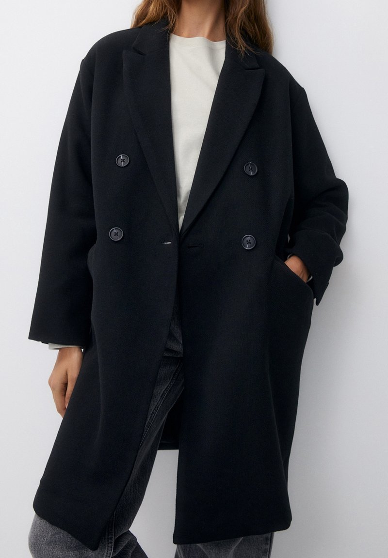 Femme portant un long manteau noir à double boutonnage sur une chemise blanche et un jean gris, une main dans la poche du manteau, debout devant un fond uni.