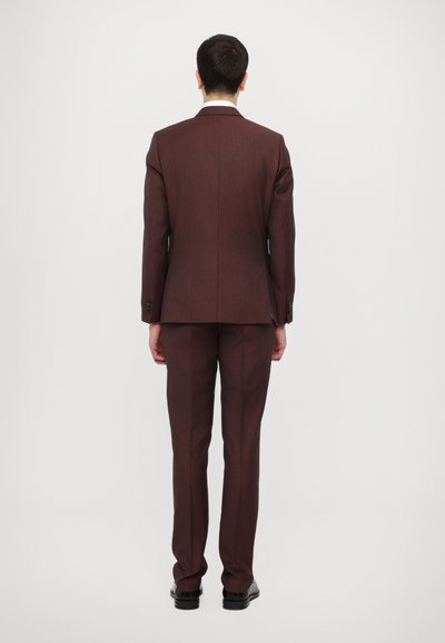 Costume marron sur mesure avec une silhouette ajustée, un tissu texturé et un éclat subtil. Comprend une fente simple et des revers standards, porté avec des chaussures noires.