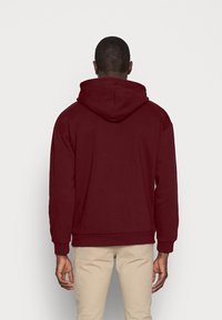 Felpa con cappuccio bordeaux dal fit rilassato, con maniche lunghe e polsini a coste, caratterizzata da una texture morbida e un design in tinta unita.
