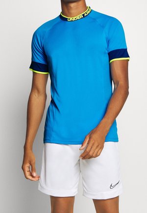 Homme portant un t-shirt de sport bleu Under Armour avec des accents néon et un short de sport blanc Nike sur un fond uni.