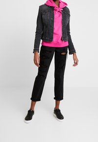 Czarna kurtka jeansowa na różowej bluzie z kapturem, zestawiona z czarnymi podartymi jeansami i czarnymi trampkami z białymi podeszwami. Codzienny strój.