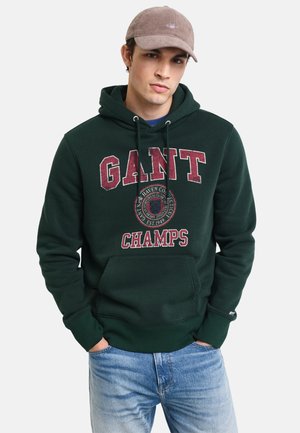 Green hooded sweatshirt featuring large "GANT CHAMPS" lettering in burgundy, front pocket, and drawstring hood. Worn with light blue jeans.