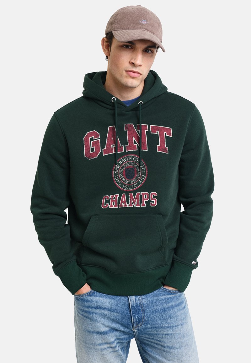 Sudadera verde con capucha que presenta grandes letras "GANT CHAMPS" en burdeos, bolsillo frontal y capucha con cordón. Combinada con jeans de color azul claro.