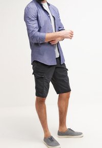 Conjunto casual de hombre que incluye una camisa de botones azul claro con mangas arremangadas, camiseta gris, pantalones cortos cargo negros y zapatos grises.