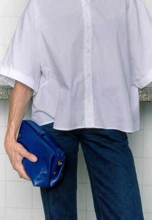 Persona che indossa una camicia bianca larga con bottoni e pantaloni scuri, che tiene una piccola borsa clutch in pelle blu contro un muro piastrellato.