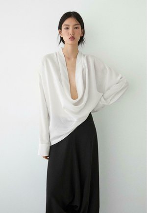 Kobieta nosząca luźną białą bluzkę z głębokim dekoltem i długą czarną spódnicę, stojąca na tle jednolitej białej ściany.