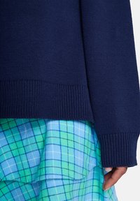 Μπλε πλεκτό πουλόβερ με ριμπ μανσέτες και τελείωμα, συνδυασμένο με φούστα σε στρώσεις με μπλε και πράσινο καρό, που διαθέτει βολάν και αντίθετα μοτίβα.