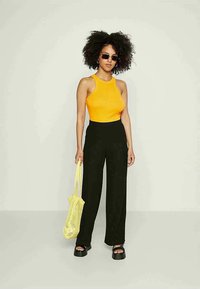 Geel fitted tanktop, zwarte wijde broek, zwarte platform sandalen en een gele mesh tote bag. Model heeft krullend haar en een zonnebril.