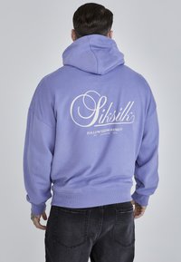 Sudadera con capucha morada y puños y dobladillo acanalados. La parte posterior presenta un gran texto de logo en blanco. Textura de tela suave, corte relajado.