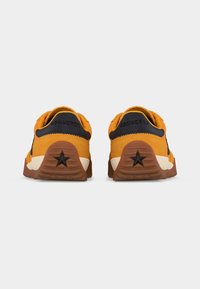 Ζευγάρι πορτοκαλί αθλητικά παπούτσια Converse, ορατά από πίσω, με μαύρο περιθώριο στη φτέρνα, λεπτομέρεια αστεριού σε καφέ σόλες και λευκές λεπτομέρειες στη μεσαία σόλα.