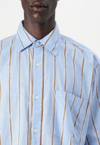Camisa azul claro con rayas verticales marrones, de mangas cortas, con botones en el frente y un bolsillo en el pecho. Fabricada con una tela texturizada.