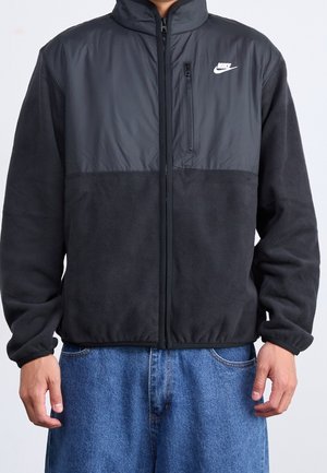 Mężczyzna ubrany w czarną polarową kurtkę Nike z zamykaną kieszenią na piersi i niebieskie dżinsy, stojący na jednolitym tle.