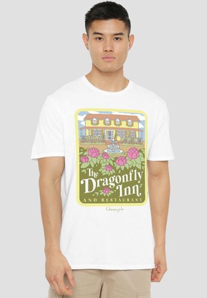 Young man wearing white T-shirt with colorful graphic of a yellow house, fountain, pink flowers, and text "The Dragonfly Inn and Restaurant."