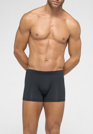 Uomo muscoloso che indossa boxer aderenti neri, in piedi con le mani dietro la schiena contro uno sfondo chiaro e uniforme.