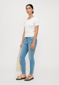 Witte kortemouwen aansluitende t-shirt, lichtblauwe skinny jeans, beige sandalen met gespen en een geweven beige handtas. Eenvoudig ontwerp, casual stijl.