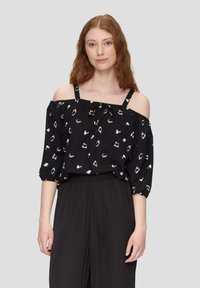 Blusa nera off-shoulder con motivi bianchi, maniche elastiche e dettaglio con laccetto al collo. Abbinata a pantaloni neri.