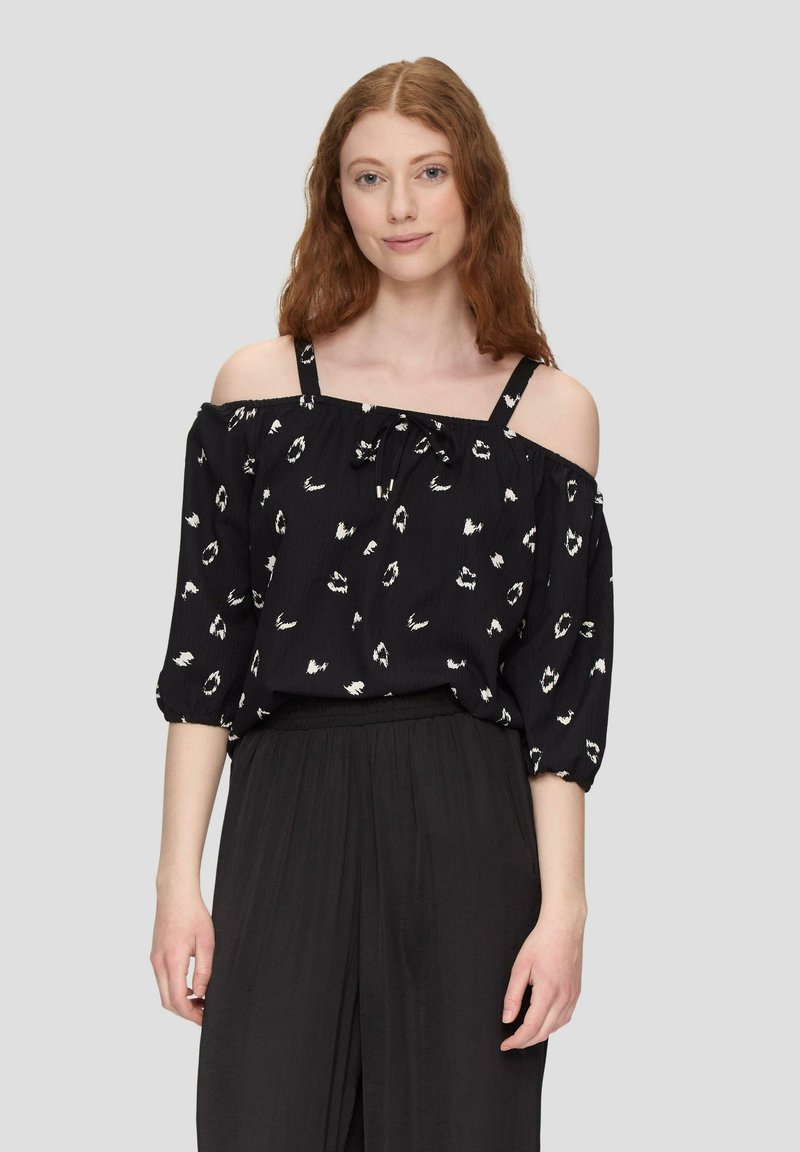 Blusa nera off-shoulder con motivi bianchi, maniche elastiche e dettaglio con laccetto al collo. Abbinata a pantaloni neri.