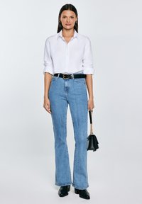 Žena s rovnými tmavými vlasy, která má na sobě bílou košili zapnutou na knoflíky, modré džíny s vysokým pasem, černý pásek, kotníkové boty a drží černou kabelku.