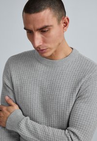 Bărbat cu păr scurt și închis la culoare, purtând un pulover texturat de culoare gri deschis, privind în jos cu o mână sprijinită pe brațul opus.