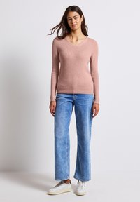 Světle růžový svetr s ribováním a výstřihem do V, dlouhými rukávy, v kombinaci se světle modrými širokými džínami a bílými teniskami.