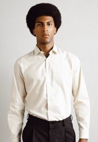 Camicia button-up color crema con maniche lunghe, abbinata a pantaloni neri. Texture liscia e design del colletto classico.