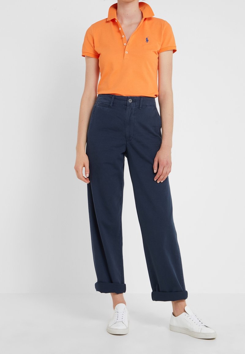 Orange pikétröja med krage, kombinerad med marinblå byxor med hög midja. Stylad med vita sneakers och en avslappnad silhuett.