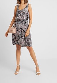 Robe noire à motif paisley, sans manches, taille cintrée et ourlet fluide. Associée à des sandales à talons blanches et un sac à main rose.