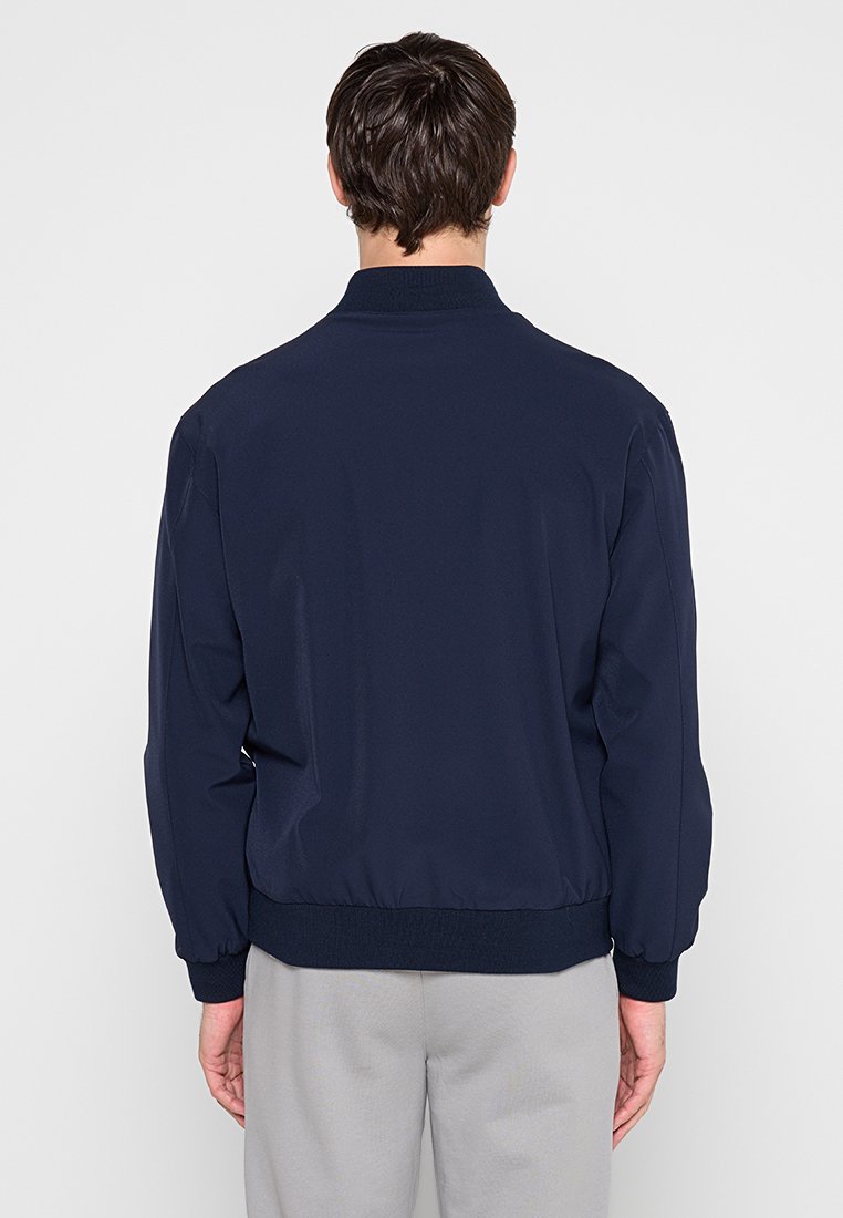 Hombre de espaldas que lleva una chaqueta bomber azul marino y pantalones gris claro frente a un fondo liso y claro.