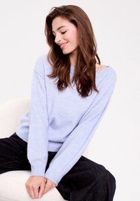 Un pull doux tricoté de couleur bleu clair avec un design décolleté et des poignets côtelés, associé à un pantalon sombre, sur un fond neutre.