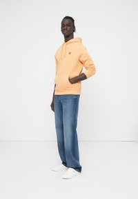 Πορτοκαλί φούτερ από βαμβάκι με τσέπη καγκουρό και κίτρινο λογότυπο, συνδυασμένο με τζιν παντελόνι ευθείας γραμμής σε ανοιχτό μπλε και λευκά αθλητικά παπούτσια.