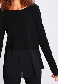 Svart långärmad topp med texturerad överdel och ett lager av genomskinlig svart nederkant. Har knapptyglar i nederkanten. Casual design.