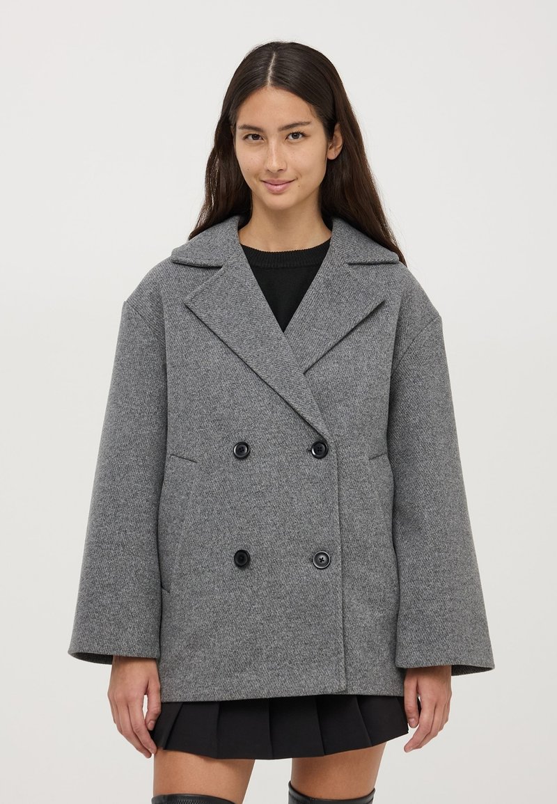 Cappotto grigio doppiopetto realizzato in tessuto testurizzato, caratterizzato da grandi bottoni neri, ampi rever e una vestibilità larga e squadrata.