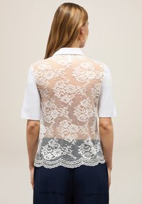 Polo blanc à manches courtes, avec un dos en dentelle transparente orné de motifs floraux et de bords festonnés, contraste avec un bas bleu marine uni.