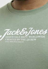 Olivgrüner Pullover mit Rundhalsausschnitt. Hat weißen Drucktext: "Jack & Jones Essentials Dept. Worldwide" und "Created by the JJ Crew."