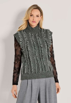 Eine blonde Frau trägt lange, schwarze Spitzenärmel unter einem strukturierten, grauen, ärmellosen Pullover und grauen, plissierten Hosen.