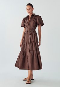 Rjava bombažna obleka s kratkimi napihnjenimi rokavi, gumbom spredaj, pasom in zasnovo z več plastmi. Obuta z črnimi trakastimi natikači.