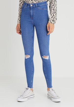 Frau trägt hoch taillierte blaue Skinny-Jeans mit zerrissenen Knien, weiße Plateausneaker und ein langärmeliges gemustertes Oberteil.
