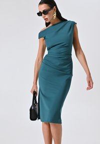 Türkisfarbenes Off-Shoulder-Kleid aus weichem Stoff, taillierte Silhouette mit drapierten Details. Model hält eine schwarze Handtasche, trägt oversized Sonnenbrillen und goldene Ohrringe.