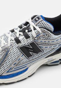 Vit och blå New Balance löparskor med svart "N"-logotyp, mesh-ovandel, vita skosnören och d�ämpad sula synlig mot en vit bakgrund.