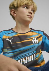 Zils un oranžs rakstains krekls ar īsām piedurknēm, ar apaļu izgriezumu, Puma logotipu un Olympique de Marseille zīmolu priekšā.