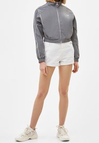 Šedá zkrácená bunda s vysokým límcem, zipovým zapínáním a bílými akcenty. Krátké bílé šortky a světlé tenisky doplňují celý outfit.