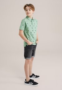 Ljusgrön polo skjorta med mörkgröna mönster, korta ärmar och vikta kragar, som är kombinerad med mörka denimshorts och svarta sneakers.