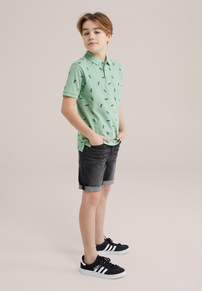 Ljusgrön polo skjorta med mörkgröna mönster, korta ärmar och vikta kragar, som är kombinerad med mörka denimshorts och svarta sneakers.