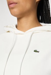 Kremfarget hettegenser laget av mykt stoff, med kengurulomme, justerbar hette med snøring, og en liten brodert grønn krokodille-logo.