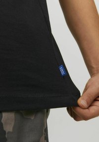 Černé bavlněné tričko s rovým lemem, s modrou značkovou etiketou na bočním švu. Textil má hladkou strukturu.