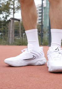 Hvide atletiske sko med en buet form og tekstureret overflade, der har en sort detalje nær snørebåndene og en mønstret sål.