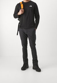 Schwarzes Langarmshirt mit kleinem Logo, kombiniert mit dunkelgrauer Hose und schwarzen Wanderschuhen. Orangener Riemen an einem schwarzen Rucksack.