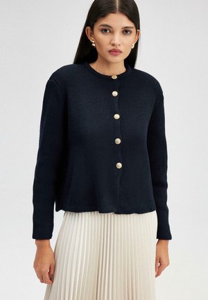 Marineblauwe gebreide cardigan met een ronde halslijn, sluiting met knopen aan de voorkant met goudkleurige knopen, en lange mouwen, gedragen over een geplisseerde rok.