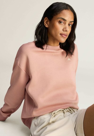 Kvinde med skulderlangt sort hår iført en lyserød sweatshirt og lyse shorts, siddende foran en ensfarvet baggrund.